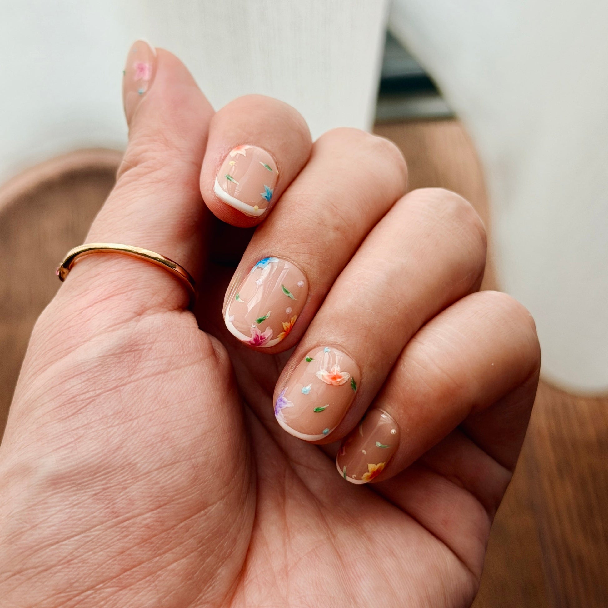 Hand with painted florals on press-on nails and a gold ring on a wooden surface