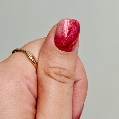 Close-up of a hand with a gold ring and a nail with red, white, and sparkly marbled polish on a gray background