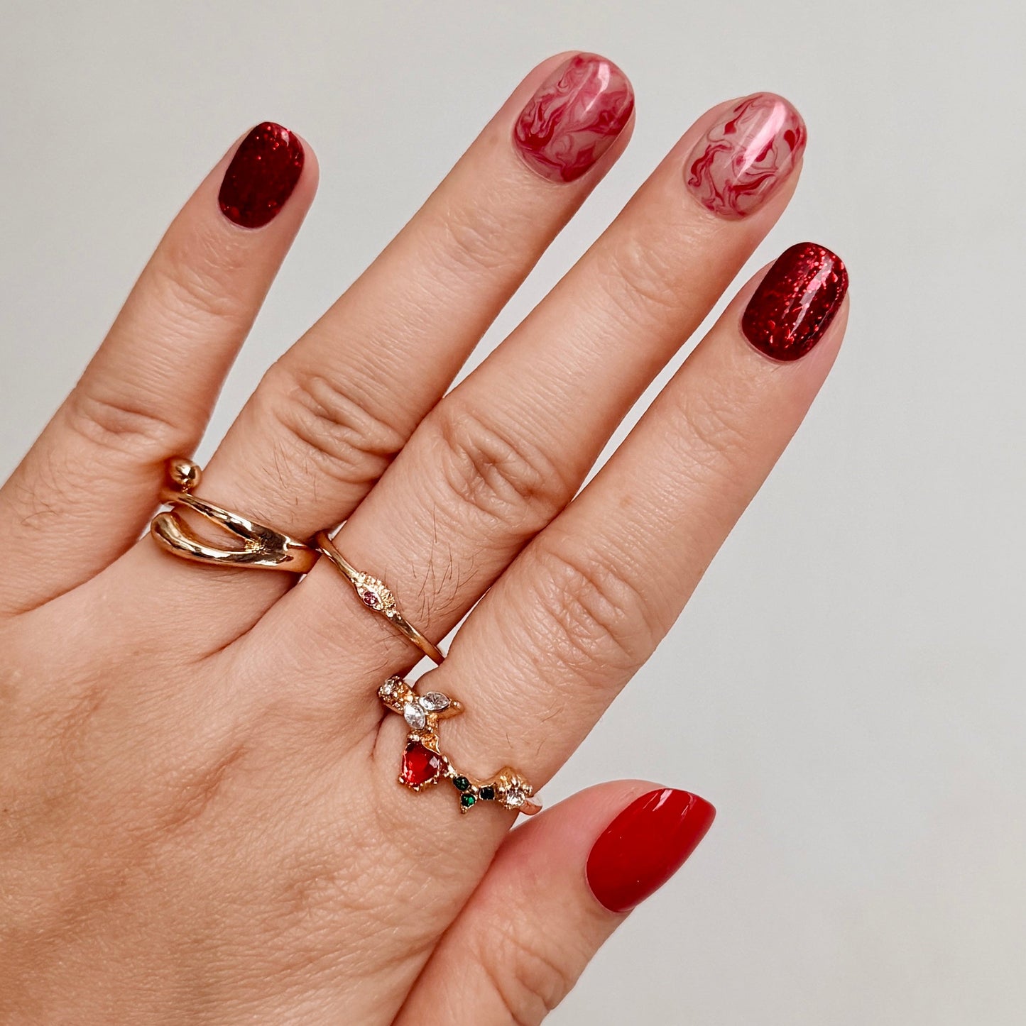 Hand with red nail polish wearing two gold rings on a neutral background