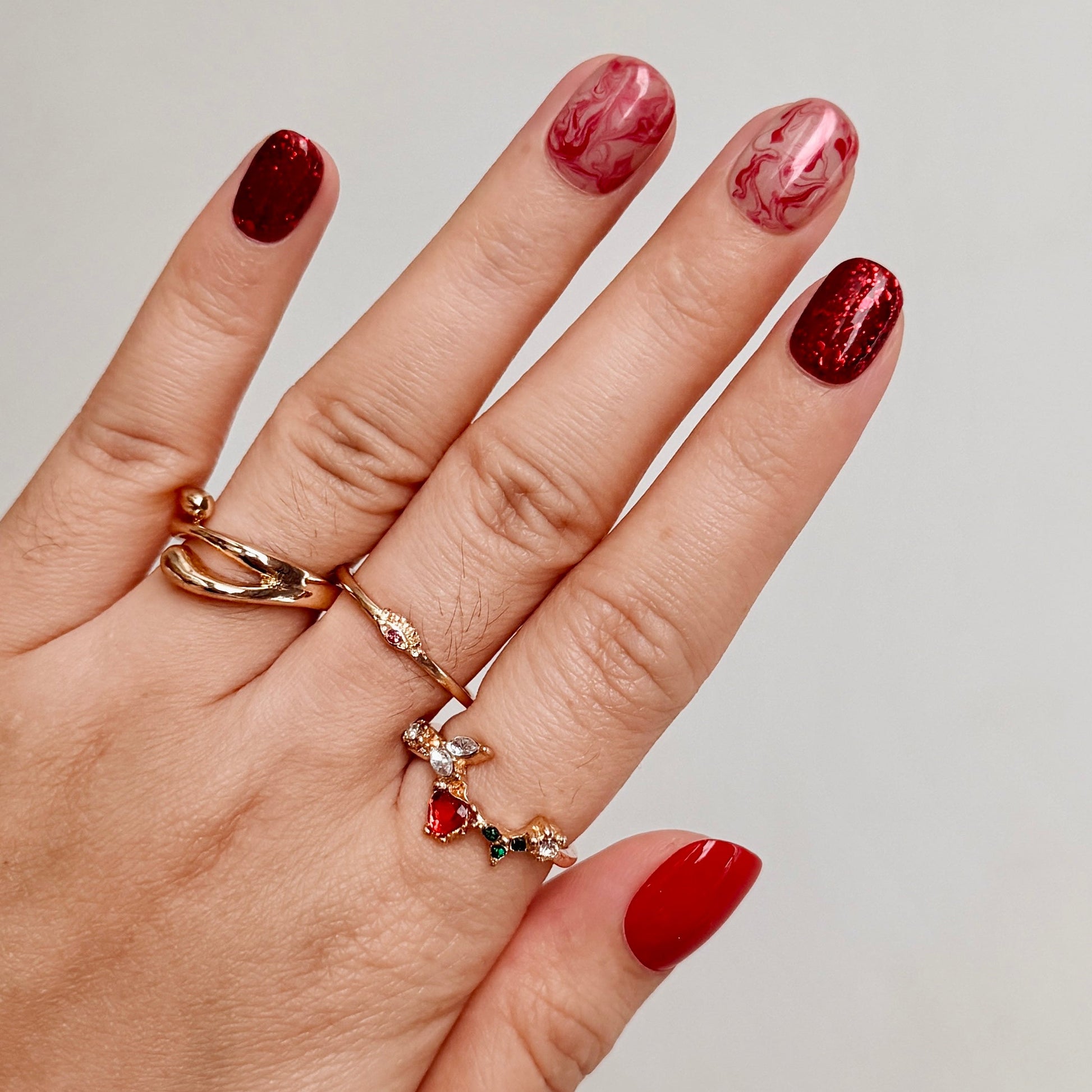 Hand with red nail polish wearing two gold rings on a neutral background